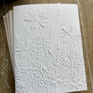 Puede incluir: Un paquete de tarjetas de felicitaci&oacute;n blancas con un dise&ntilde;o en relieve de varias flores silvestres, incluyendo margaritas, helechos y hierbas altas. La textura crea un efecto tridimensional en la superficie del papel, resaltando los intrincados detalles bot&aacute;nicos del arreglo floral en un acabado blanco monocrom&aacute;tico.