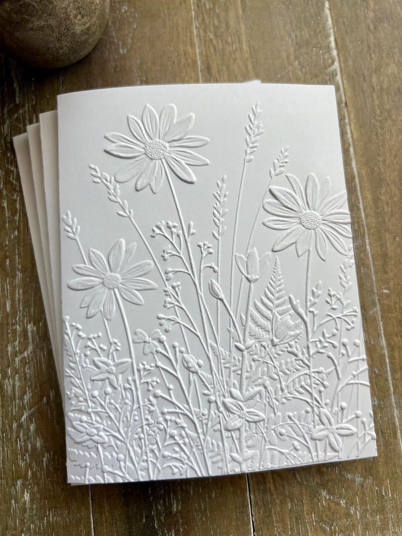 Puede incluir: Un paquete de tarjetas de felicitaci&oacute;n blancas con un dise&ntilde;o en relieve de varias flores silvestres, incluyendo margaritas, helechos y hierbas altas. La textura crea un efecto tridimensional en la superficie del papel, resaltando los intrincados detalles bot&aacute;nicos del arreglo floral en un acabado blanco monocrom&aacute;tico.