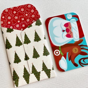 May include: A Christmas-themed gift card and envelope. The gift card features Santa Claus riding a reindeer against a blue background. The envelope has a pattern of green Christmas trees on a cream background, with a red interior decorated with white snowflakes.
