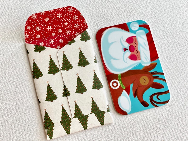 May include: A Christmas-themed gift card and envelope. The gift card features Santa Claus riding a reindeer against a blue background. The envelope has a pattern of green Christmas trees on a cream background, with a red interior decorated with white snowflakes.