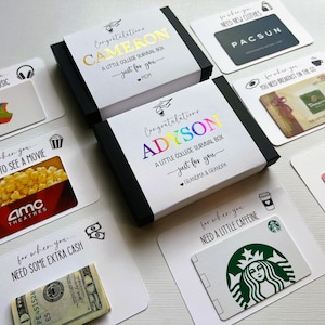 Op de afbeelding: Twee zwarte geschenkdozen met witte hoezen met de tekst Congratulations CAMERON A LITTLE COLLEGE SURVIVAL BOX en Congratulations ADYSON A LITTLE COLLEGE SURVIVAL BOX, omringd door cadeaubonnen voor Apple, PacSun, Panera, AMC, Crumbl en Starbucks.