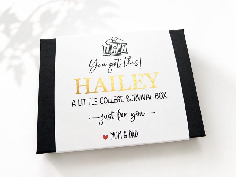 Op de afbeelding: Een rechthoekige zwarte geschenkdoos met een witte papieren wikkel met een zwarte lijntekening van een schoolgebouw, de tekst "You got this! HAILEY A LITTLE COLLEGE SURVIVAL BOX just for you" in goud en zwart, en een rood hartje.