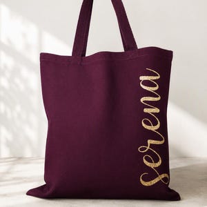 May include: A deep burgundy canvas tote bag featuring two matching fabric handles and the name Serena printed vertically along the right side in a shimmering gold glitter script font. The bag has a rectangular shape and a solid dark fabric.