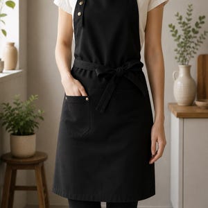 May include: A solid black apron featuring a neck strap, a waist tie, and a single front pocket. The apron includes metallic hardware details on the shoulder strap and pocket corner, providing a functional design for kitchen or utility use.