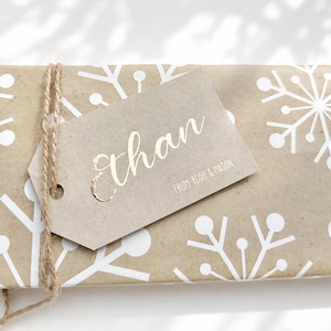 May include: A rectangular gift wrapped in brown paper with a white snowflake pattern, tied with natural jute twine. A light brown cardstock gift tag is attached, featuring the name Ethan in gold foil script and the text FROM ADDY & MASON.