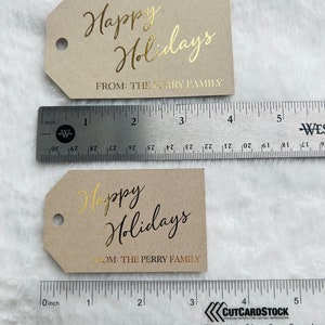 May include: Two rectangular kraft paper gift tags with angled corners and a circular hole. The top tag is 4 inches wide with gold foil text reading Happy Holidays FROM: THE PERRY FAMILY. The bottom tag is 3 inches wide with black text.
