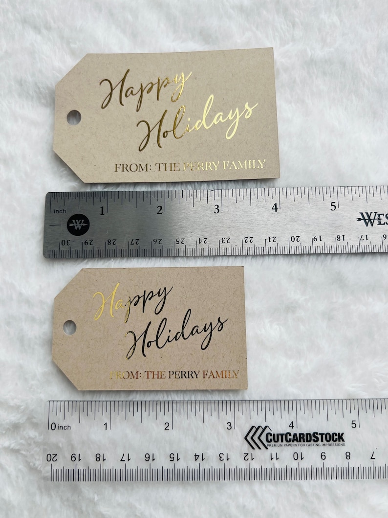 May include: Two rectangular kraft paper gift tags with angled corners and a circular hole. The top tag is 4 inches wide with gold foil text reading Happy Holidays FROM: THE PERRY FAMILY. The bottom tag is 3 inches wide with black text.
