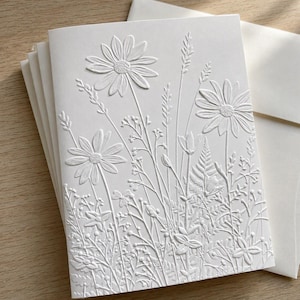 May include: A white greeting card featuring a raised, embossed design of various wildflowers and grasses. The texture creates a monochromatic floral pattern with daisies, ferns, and delicate stems across the front surface of the folded paper card.