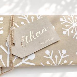May include: A rectangular gift wrapped in light brown paper with a white snowflake pattern, tied with natural jute twine. A light brown paper gift tag is attached, featuring the name Ethan in gold foil and the text FROM ADDY & MASON.