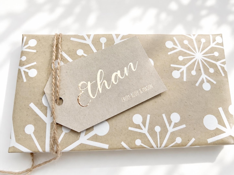 May include: A rectangular gift wrapped in light brown paper with a white snowflake pattern, tied with natural jute twine. A light brown paper gift tag is attached, featuring the name Ethan in gold foil and the text FROM ADDY & MASON.