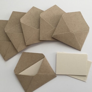 Puede incluir: Un conjunto de seis sobres peque&ntilde;os de papel kraft marr&oacute;n con solapas puntiagudas, dispuestos en semic&iacute;rculo, con un sobre abierto que revela una tarjeta de color crema lisa en su interior, acompa&ntilde;ado de una peque&ntilde;a pila de tarjetas rectangulares a juego.