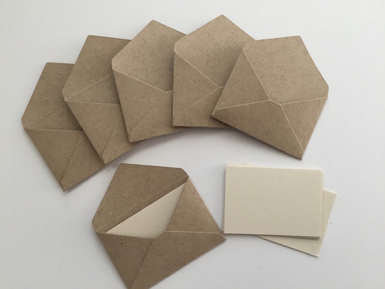 Puede incluir: Un conjunto de seis sobres peque&ntilde;os de papel kraft marr&oacute;n con solapas puntiagudas, dispuestos en semic&iacute;rculo, con un sobre abierto que revela una tarjeta de color crema lisa en su interior, acompa&ntilde;ado de una peque&ntilde;a pila de tarjetas rectangulares a juego.