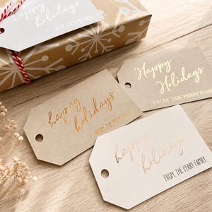 May include: Three rectangular gift tags with angled corners, each featuring the text "Happy Holidays" in cursive and "FROM: THE PERRY FAMILY" in block letters. The tags are in tan, light brown, and off-white, with metallic foil lettering.