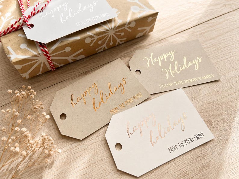 May include: Three rectangular gift tags with angled corners, each featuring the text "Happy Holidays" in cursive and "FROM: THE PERRY FAMILY" in block letters. The tags are in tan, light brown, and off-white, with metallic foil lettering.