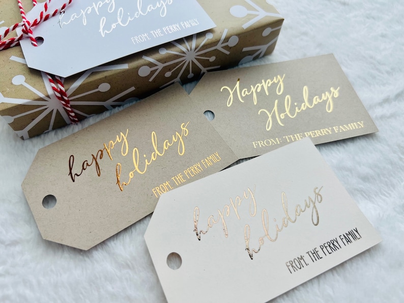 May include: Three rectangular gift tags with angled corners and a circular hole, featuring the gold foil text "Happy Holidays" and "FROM: THE PERRY FAMILY" in a script and sans-serif font, displayed alongside a gift box wrapped in snowflake paper.
