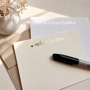 May include: A set of cream and white stationery cards featuring the name melissa brooks in gold foil lettering with small heart icons. A brown kraft paper envelope is tucked underneath the cards, and a black Sharpie marker rests on top.