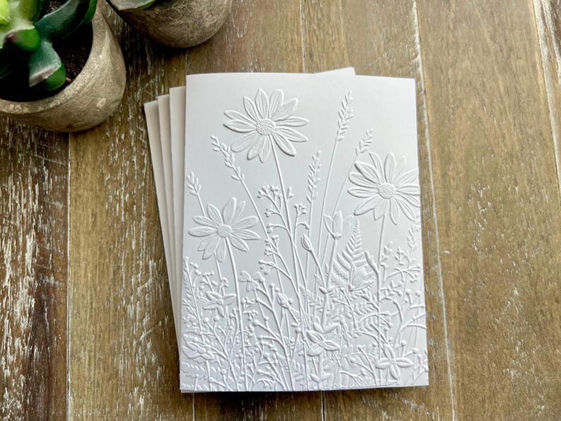 Puede incluir: Un mont&oacute;n de tarjetas de felicitaci&oacute;n blancas con un dise&ntilde;o en relieve de varias flores silvestres, margaritas y helechos. La textura crea un sutil patr&oacute;n floral monocrom&aacute;tico en la parte frontal de las tarjetas, resaltando los detalles bot&aacute;nicos.