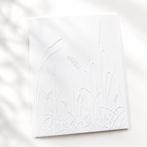 Op de afbeelding: Een rechthoekige witte wenskaart met een verhoogd, reli&euml;f ontwerp van diverse wilde grassen en tarwestengels. De monochrome textuur cre&euml;ert een subtiel reli&euml;feffect op het oppervlak van het papier, wat de botanische vormen benadrukt.