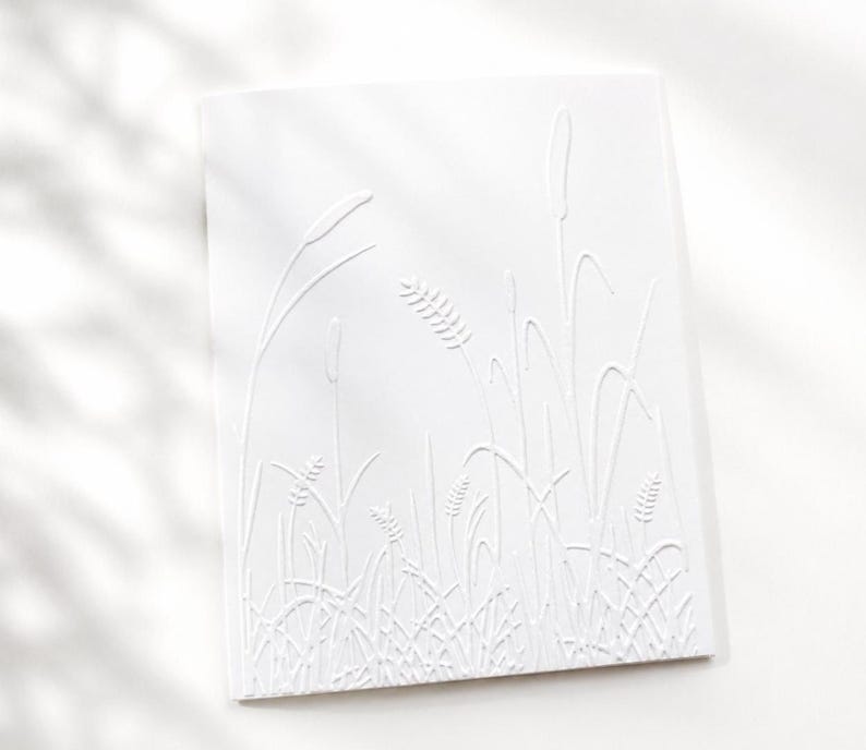 Op de afbeelding: Een rechthoekige witte wenskaart met een verhoogd, reli&euml;f ontwerp van diverse wilde grassen en tarwestengels. De monochrome textuur cre&euml;ert een subtiel reli&euml;feffect op het oppervlak van het papier, wat de botanische vormen benadrukt.