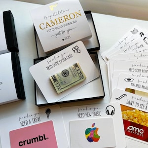 Op de afbeelding: Een verzameling witte geschenkdozen en kaarten voor een college survivalpakket. De dozen bevatten zwarte en gouden tekst met namen zoals Cameron en Emily. De kaarten tonen zinnen zoals Need Some Extra Cash met een biljet van twintig dollar.