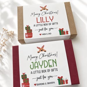 May include: Two rectangular gift boxes with white paper sleeves. The top box is brown with text reading Merry Christmas! LILLY A LITTLE BOX OF GIFTS just for you MOM & DAD. The bottom box is maroon with text reading Merry Christmas! JAYDEN A LITTLE BOX OF GIFTS just for you GRANDMA & GRANDPA. Both feature reindeer and gift illustrations.