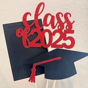 May include: A graduation-themed cake topper with a red glitter "Class of 2025" design. The topper includes a black graduation cap with a red tassel and a black diploma. The design is mounted on wooden sticks.