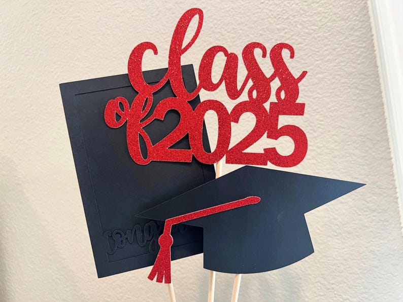 May include: A graduation-themed cake topper with a red glitter "Class of 2025" design. The topper includes a black graduation cap with a red tassel and a black diploma. The design is mounted on wooden sticks.