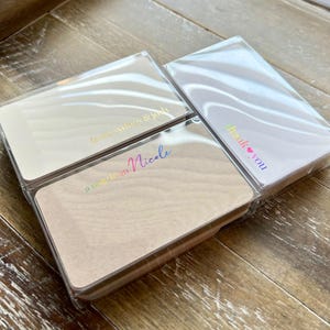 May include: Three stacks of rectangular cards in clear plastic packaging. The cards have a light wood grain appearance and feature iridescent text that reads "thank you", "a baptism by Nicole", and "thessalonike & josh". The cards are on a wooden surface.