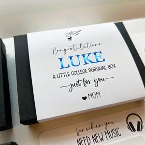 Op de afbeelding: Een zwarte rechthoekige geschenkdoos met een witte papieren hoes met een icoon van een afstudeerhoed en de tekst Congratulations LUKE A LITTLE COLLEGE SURVIVAL BOX just for you MOM, waarbij de naam LUKE in blauwe metallic folie is gedrukt.