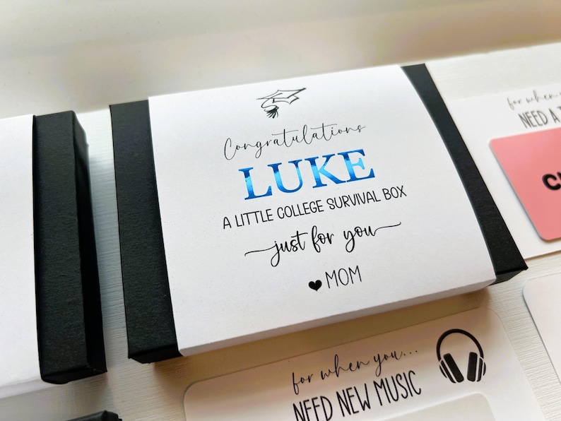 Op de afbeelding: Een zwarte rechthoekige geschenkdoos met een witte papieren hoes met een icoon van een afstudeerhoed en de tekst Congratulations LUKE A LITTLE COLLEGE SURVIVAL BOX just for you MOM, waarbij de naam LUKE in blauwe metallic folie is gedrukt.