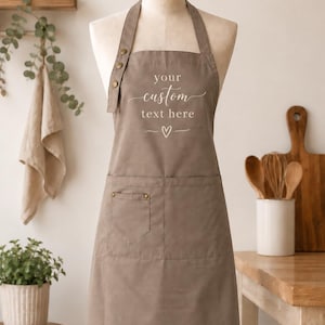 May include: A taupe apron featuring a front pocket and adjustable neck strap with three brass-colored buttons. The chest area displays the white text "your custom text here" above a small heart symbol and two horizontal lines, centered on the fabric.