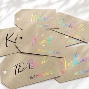 May include: A collection of rectangular kraft paper gift tags with rounded corners and a punched hole. Each tag features custom names and the text "TORIE'S BACHELORETTE 2025" printed in either solid black or a reflective, iridescent rainbow foil finish.