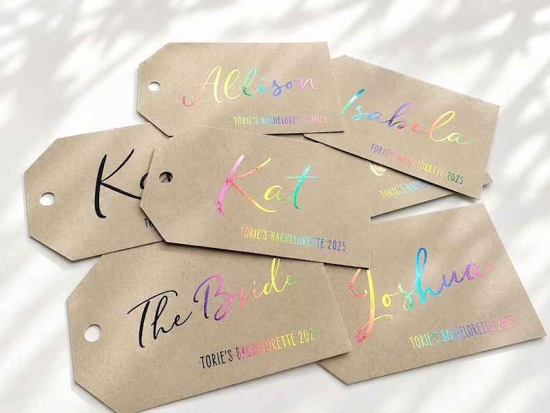 May include: A collection of rectangular kraft paper gift tags with rounded corners and a punched hole. Each tag features custom names and the text "TORIE'S BACHELORETTE 2025" printed in either solid black or a reflective, iridescent rainbow foil finish.