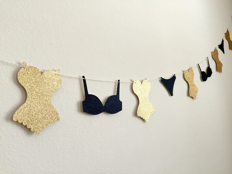 May include: A gold glitter and black paper garland with various lingerie shapes, including bras, corsets, and underwear.
