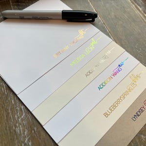 May include: A stack of rectangular paper cards featuring various names and initials in metallic foil lettering, including BRITTANY THOMAS, MELISSA LEON, ADDISON WANG, BLUEBERRYSPRINKLES, and LYNDSEY SMITH, accompanied by a black marker pen resting on top.