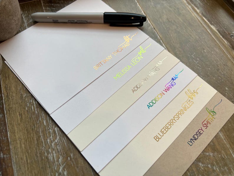May include: A stack of rectangular paper cards featuring various names and initials in metallic foil lettering, including BRITTANY THOMAS, MELISSA LEON, ADDISON WANG, BLUEBERRYSPRINKLES, and LYNDSEY SMITH, accompanied by a black marker pen resting on top.