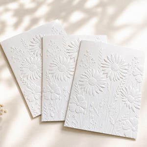 May include: Three white rectangular greeting cards featuring a raised, embossed floral design. The texture depicts various wildflowers, including daisies and tall grasses, creating a monochromatic, tactile surface across the front of each card.