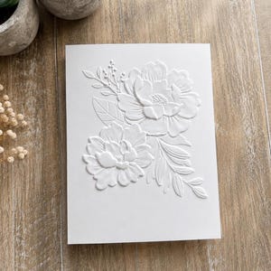 May include: A rectangular white card featuring a raised, embossed floral design on the front. The embossed pattern displays two large, detailed peony-like flowers surrounded by smaller leaves, stems, and buds, creating a textured, monochromatic look.