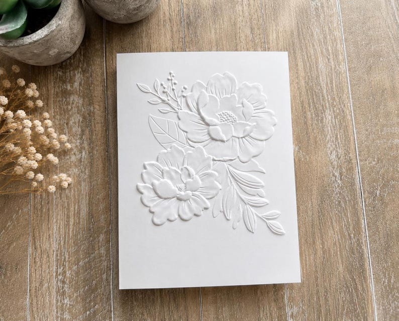 May include: A rectangular white card featuring a raised, embossed floral design on the front. The embossed pattern displays two large, detailed peony-like flowers surrounded by smaller leaves, stems, and buds, creating a textured, monochromatic look.