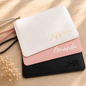 May include: Three rectangular wristlet pouches in white, light pink, and black. The white pouch features the gold text Tiffany, the pink pouch features the white text Amanda, and the black pouch features the black text JHR. Each has a wrist strap.