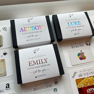 Op de afbeelding: Een verzameling witte rechthoekige geschenkdozen en kaarten voor studenten. De dozen zijn voorzien van gepersonaliseerde namen in metallic folie, afstudeerhoed-iconen en de tekst A Little College Survival Box. Bijbehorende kaarten tonen cadeaubonnen voor Panera, Apple en AMC, samen met een biljet van twintig dollar, elk gelabeld met specifieke behoeften zoals ontbijt, muziek of films.