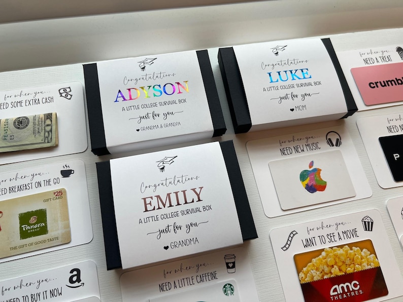 Op de afbeelding: Een verzameling witte rechthoekige geschenkdozen en kaarten voor studenten. De dozen zijn voorzien van gepersonaliseerde namen in metallic folie, afstudeerhoed-iconen en de tekst A Little College Survival Box. Bijbehorende kaarten tonen cadeaubonnen voor Panera, Apple en AMC, samen met een biljet van twintig dollar, elk gelabeld met specifieke behoeften zoals ontbijt, muziek of films.