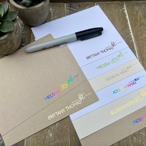 May include: A black marker resting on a stack of stationery featuring personalized names and initials in various metallic and rainbow foil finishes, including Brittany Thomas, Melissa Leon, Addison Wang, Blueberrysprinkles, and Lyndsey Smith.