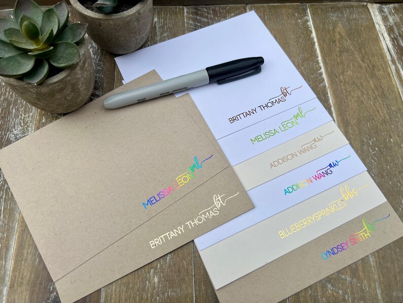 May include: A black marker resting on a stack of stationery featuring personalized names and initials in various metallic and rainbow foil finishes, including Brittany Thomas, Melissa Leon, Addison Wang, Blueberrysprinkles, and Lyndsey Smith.