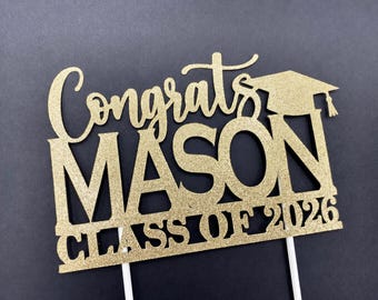 May include: A gold glitter cake topper featuring the text Congrats MASON CLASS OF 2026 with a graduation cap icon. The design is mounted on two white sticks, displaying the name Mason and the graduation year in a bold, decorative font style.
