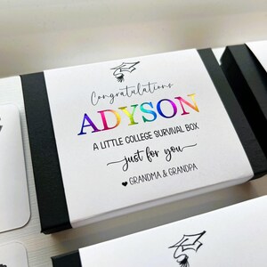 Op de afbeelding: Een rechthoekige zwart-witte geschenkdoos met een wit label voorzien van een afstudeerhoed-icoon en de tekst Congratulations ADYSON A LITTLE COLLEGE SURVIVAL BOX just for you GRANDMA & GRANDPA. De naam ADYSON is in een regenboogfolie afwerking.