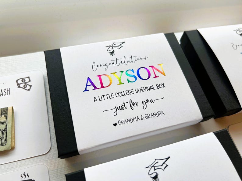 Op de afbeelding: Een rechthoekige zwart-witte geschenkdoos met een wit label voorzien van een afstudeerhoed-icoon en de tekst Congratulations ADYSON A LITTLE COLLEGE SURVIVAL BOX just for you GRANDMA & GRANDPA. De naam ADYSON is in een regenboogfolie afwerking.
