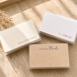 May include: Three clear plastic cases containing stacks of rectangular note cards. One stack is cream with the text from sydney & josh, one is white with the text thank you in rainbow gradient, and one is kraft brown with a note from Nicole.