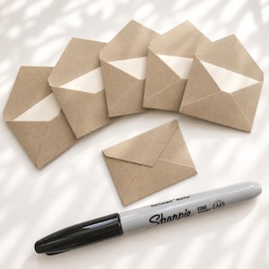 May include: Six small brown kraft paper envelopes with triangular flaps, five of which are open and one is closed, arranged near a grey Sharpie fine point permanent marker with black text and a black cap on a white surface.