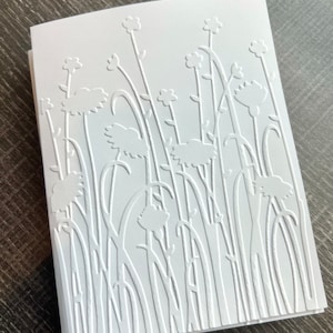 May include: A stack of white greeting cards with an embossed floral design. The design features stylized flowers and tall grass-like stems. The cards are made of thick, textured paper.